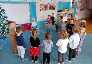 Dzieci stoją w kole na niebieskim dywanie. Na środku koła siedzi nasz solenizant z koroną na głowie.