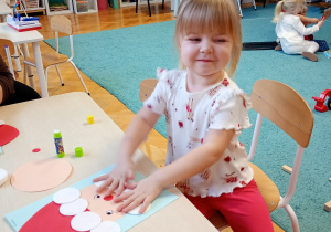 Rozalka w białej bluzce i czerwonych spodniach siedzi przy stoliku. Na stoliku leży jej gotowa praca Mikołaja