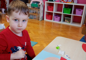 Bartuś w czerwonej bluzce siedzi przy stoliku. Przed nim leży rozpoczęta praca, którą starannie ozdabia, rozwijając swoją kreatywność i zdolności manualne