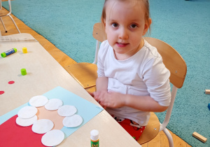 Ala w białej bluzce siedzi przy stoliku. Przed nią leży rozpoczęta praca, którą starannie ozdabia, rozwijając swoją kreatywność i zdolności manualne