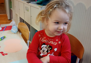 Oliwka w czerwonej sukience siedzi przy stoliku. Przed nią leży rozpoczęta praca, którą starannie ozdabia, rozwijając swoją kreatywność i zdolności manualne