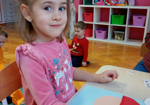 Milana w różowej bluzce siedzi przy stoliku. Przed nią leży rozpoczęta praca, którą starannie ozdabia, rozwijając swoją kreatywność i zdolności manualne