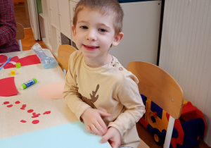 Oluś siedzi przy stoliku. Przed nim niebieska kartka, na której będzie wykonywał pracę plastyczną