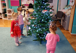 Dzieci z radością przywieszają bombki na choince, która stoi na niebieskim dywanie. Na czubku choinki znajduje się gwiazdka, a całość rozświetlają świecące lampki