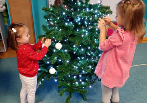 Dzieci z radością przywieszają bombki na choince, która stoi na niebieskim dywanie. Na czubku choinki znajduje się gwiazdka, a całość rozświetlają świecące lampki