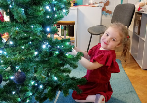 Telimena z radością przywiesza bombki na choince, która stoi na niebieskim dywanie. Całość rozświetlają świecące lampki