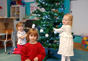 Dzieci z radością przywieszają bombki na choince, która stoi na niebieskim dywanie. Na czubku choinki znajduje się gwiazdka, a całość rozświetlają świecące lampki