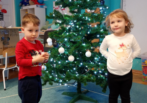 Dzieci z radością przywieszają bombki na choince, która stoi na niebieskim dywanie. Na czubku choinki znajduje się gwiazdka, a całość rozświetlają świecące lampki