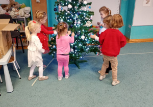 Dzieci z radością przywieszają bombki na choince, która stoi na niebieskim dywanie. Na czubku choinki znajduje się gwiazdka, a całość rozświetlają świecące lampki