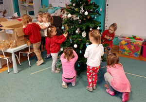 Dzieci z radością przywieszają bombki na choince, która stoi na niebieskim dywanie. Na czubku choinki znajduje się gwiazdka, a całość rozświetlają świecące lampki