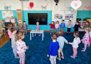 Dzieci stoją w kole na niebieskim dywanie. Na środku koła siedzu solenizantka w koronie. Przed Julką na niebieskim stoliku stoi kolorowy tort