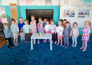 Dzieci stoją na niebieskim dywanie. Na środku siedzu solenizantka w koronie. Przed Julką na niebieskim stoliku stoi kolorowy tort