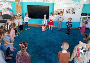 Dzieci stoją w kole na niebieskim dywanie. Na środku siedzi Jaś z koroną na głowie. Trzyma kolorową torebkę ze słodkościami.