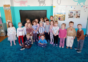 Dzieci stoją na niebieskim dywanie, na środku, na krzesle siedzi Jaś z koroną na głowie.