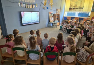 Dzieci oglądają bajkę na drewnianych krzesełkach