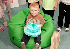 Wiktor siedzi na pufie z tortem z papieru, dzieci śpiewają mu 100 lat.
